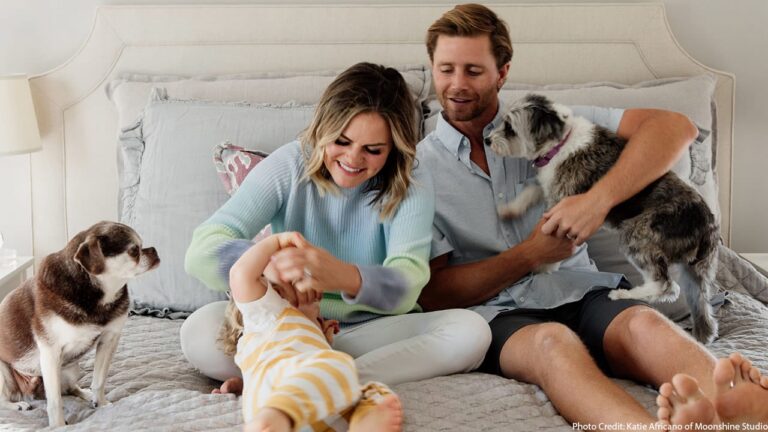 Couple and their two dogs play with a baby on a bed smiling in a cozy bedroom scene